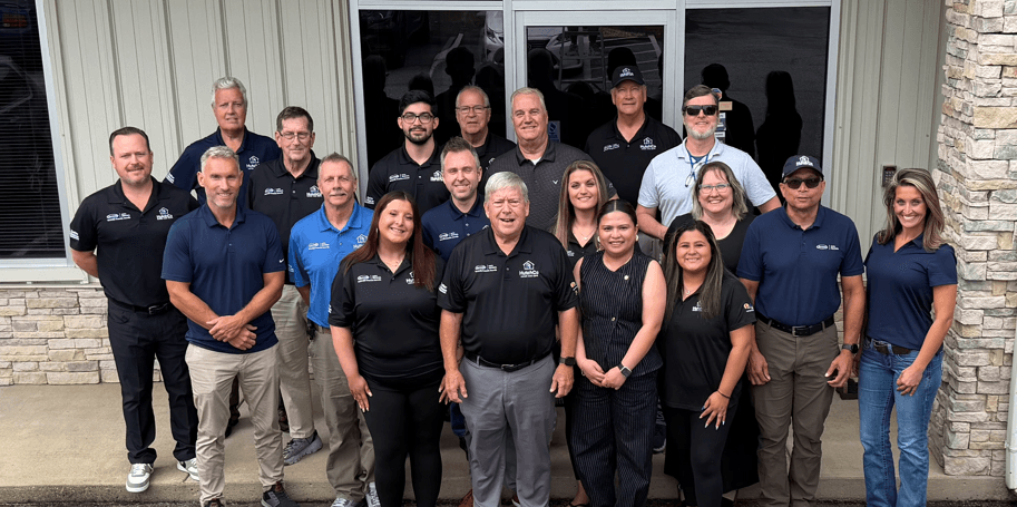 Group photo of approximately 20 team members wearing matching company polo shirts, posing together in front of a building with brick and metal siding