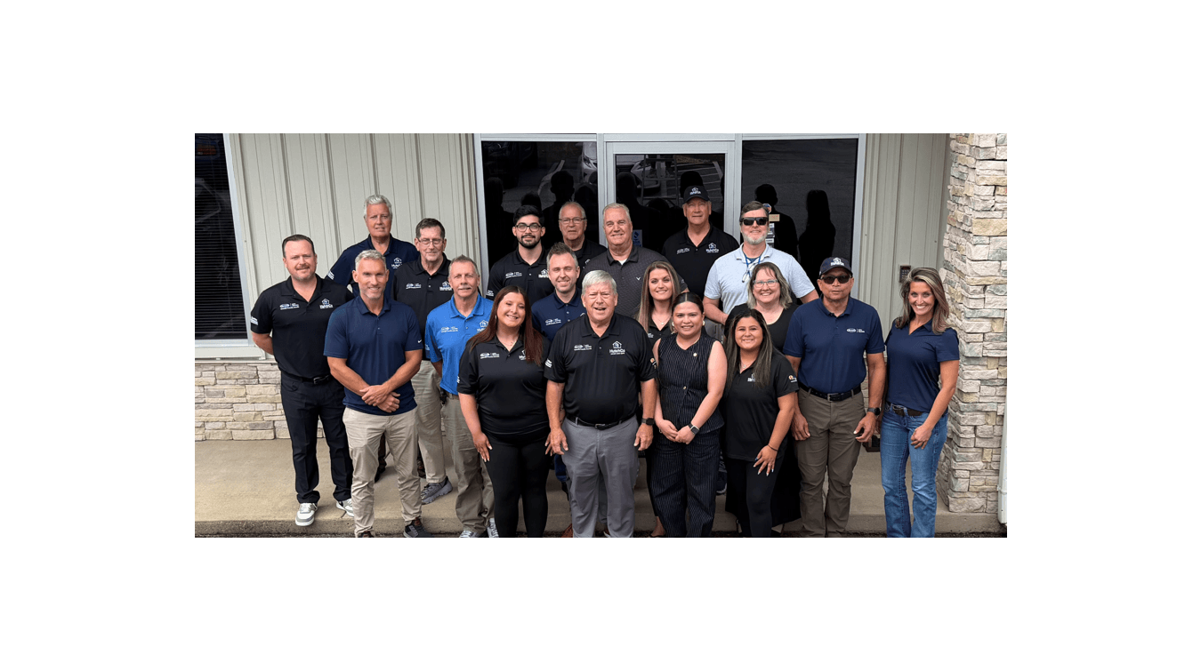 Group photo of approximately 20 team members wearing matching company polo shirts, posing together in front of a building with brick and metal siding